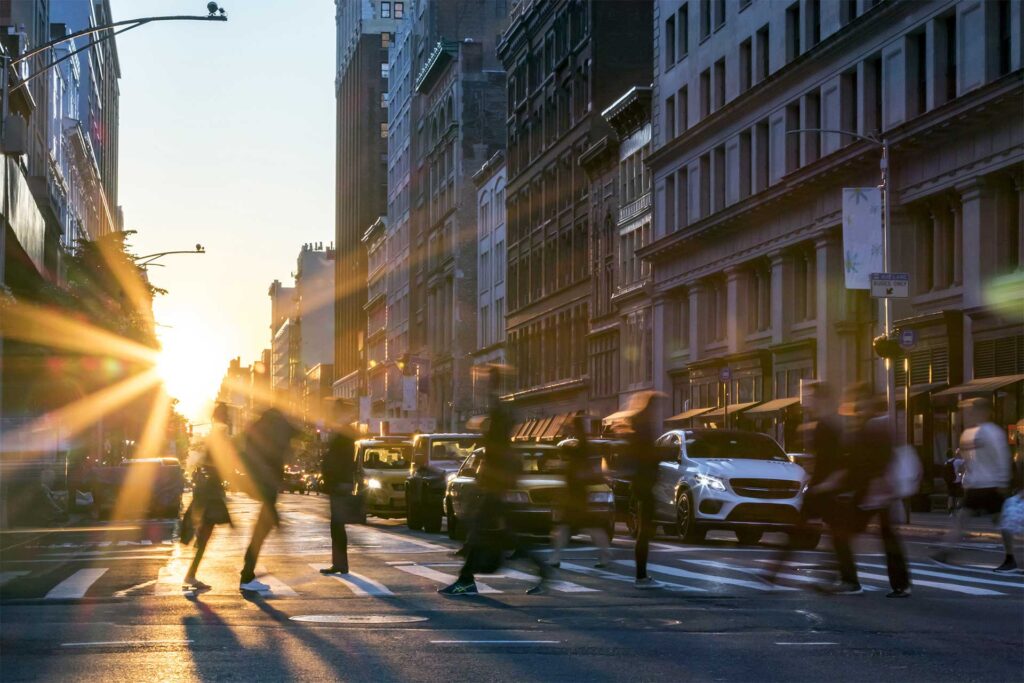 people walking across crosswalk in new york city with sunrise between buildings